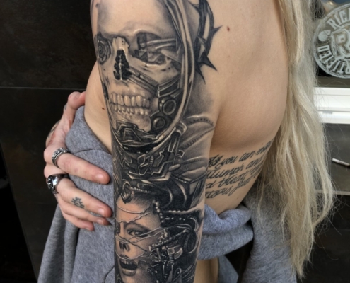 Tatuaje en el brazo de una mujer de una calavera con corona de espinas y un rostro de mujer terrorífico con detalles biomechanicos.