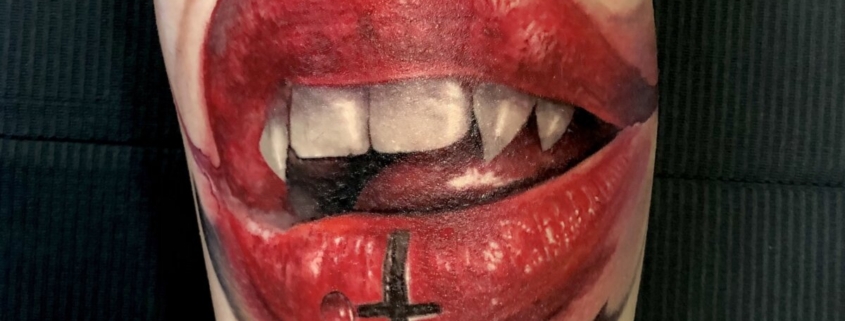 Tatuaje en el muslo de una boca de vampira sexy con una cruz invertida en el labio inferior.