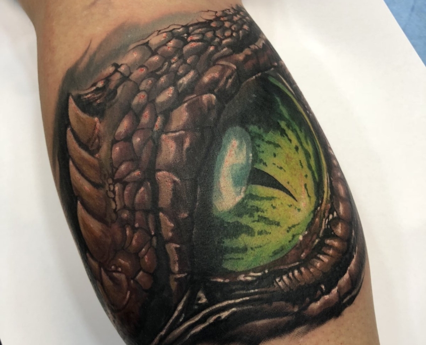 Tatuaje grande de un ojo de dragon de color verde en el gemelo de un hombre.